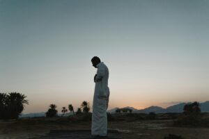 Silhouette of a man in traditional clothing praying outdoors at dawn.